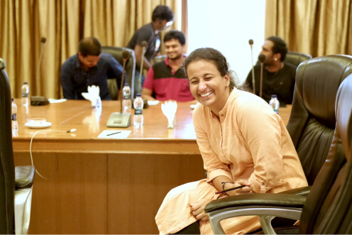 A smiling Tarkan (Vidhya) sitting at a conference table with other Tarkans talking in the background